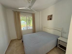 A bedroom with a bed, a ceiling fan, a window with curtains, a wall-mounted picture, and a bedside table.