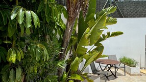A patio area with a table and chairs, surrounded by lush greenery and a white wall.