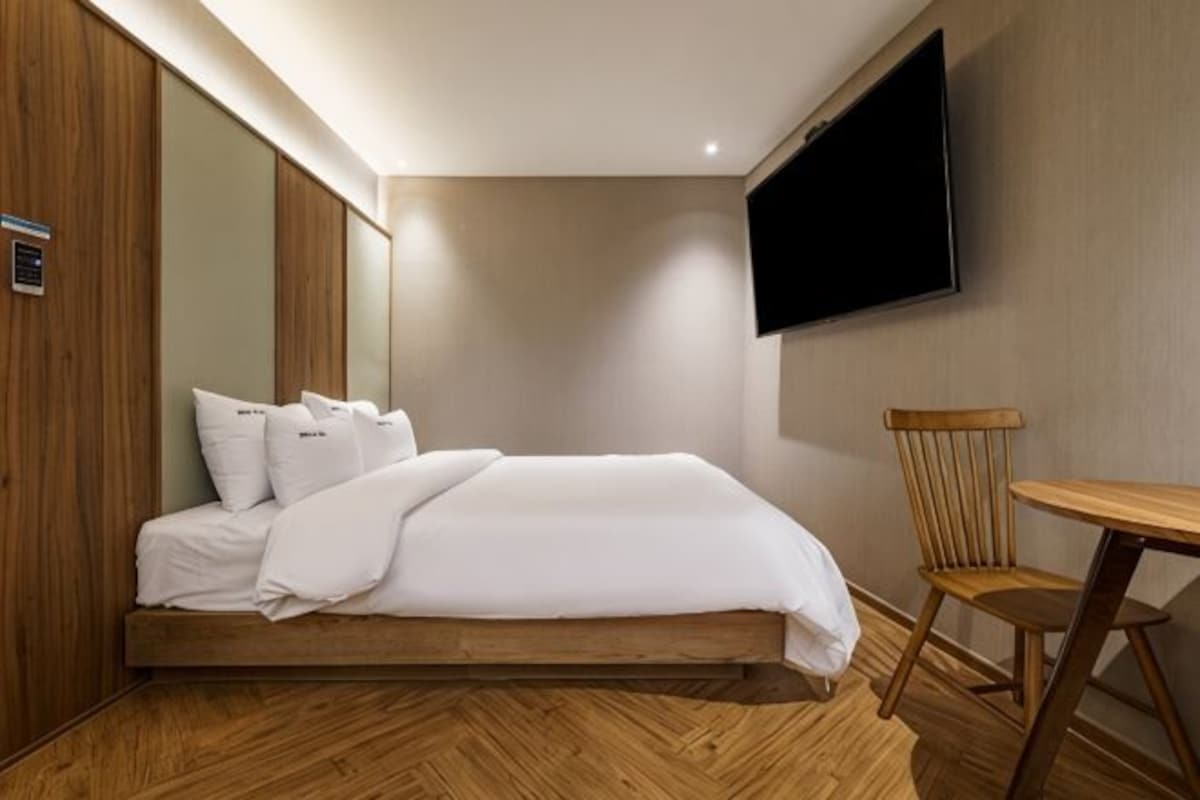 A neatly made bed with pillows, a wooden floor, a TV mounted on the wall, and a small wooden table.