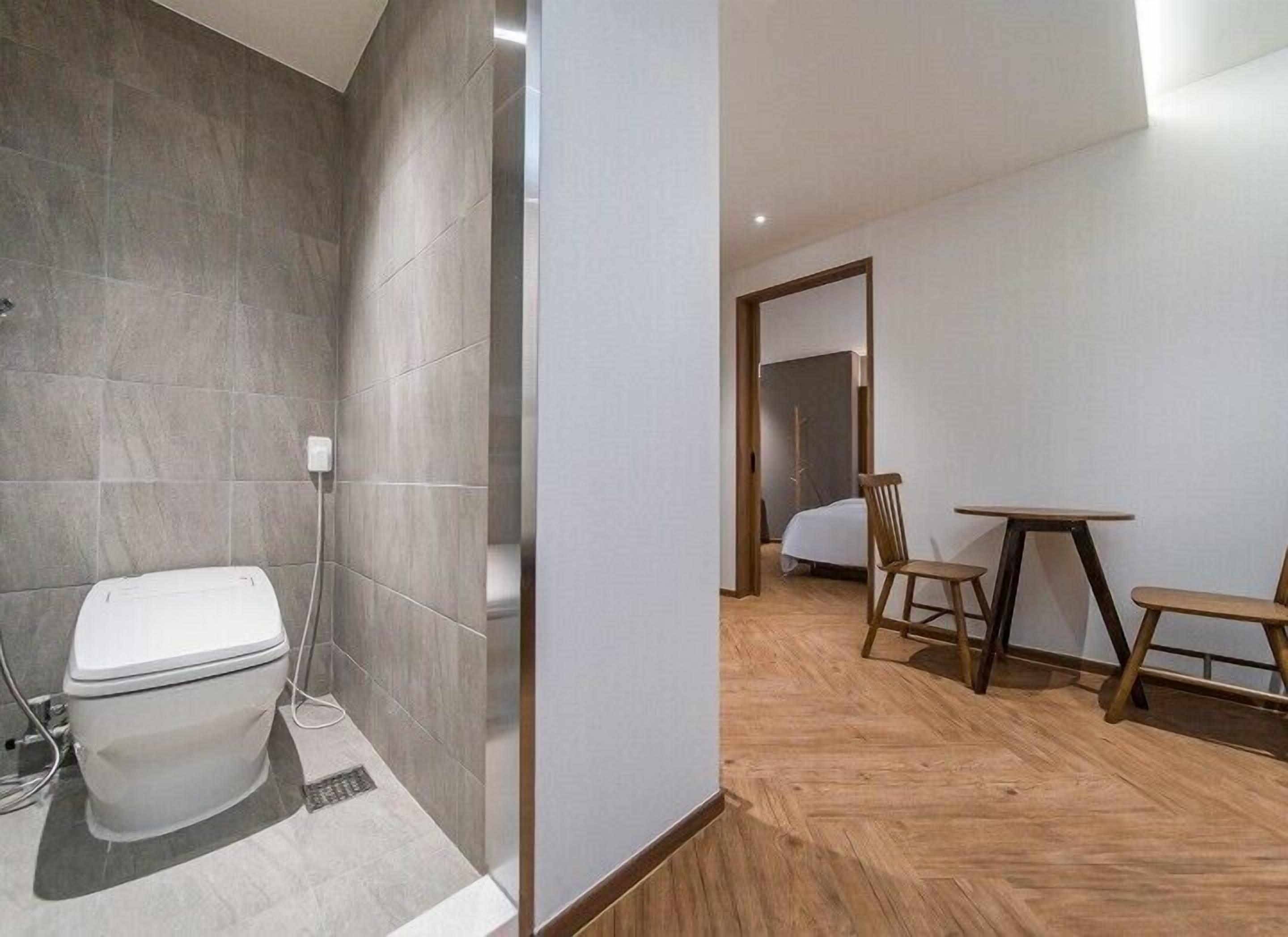 A bathroom with a toilet and a shower area, and a bedroom with a bed, a wooden table, and two chairs.