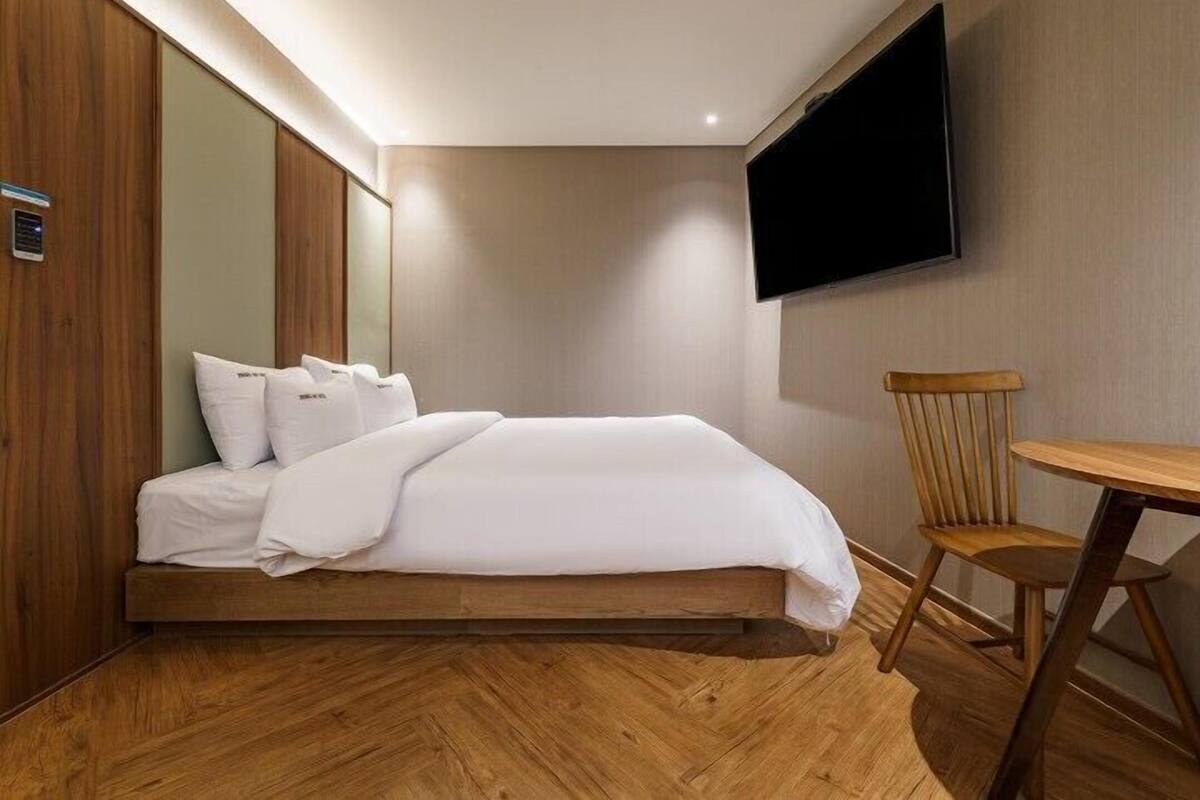 A modern hotel room with a bed, wooden flooring, a TV mounted on the wall, and a small wooden chair.