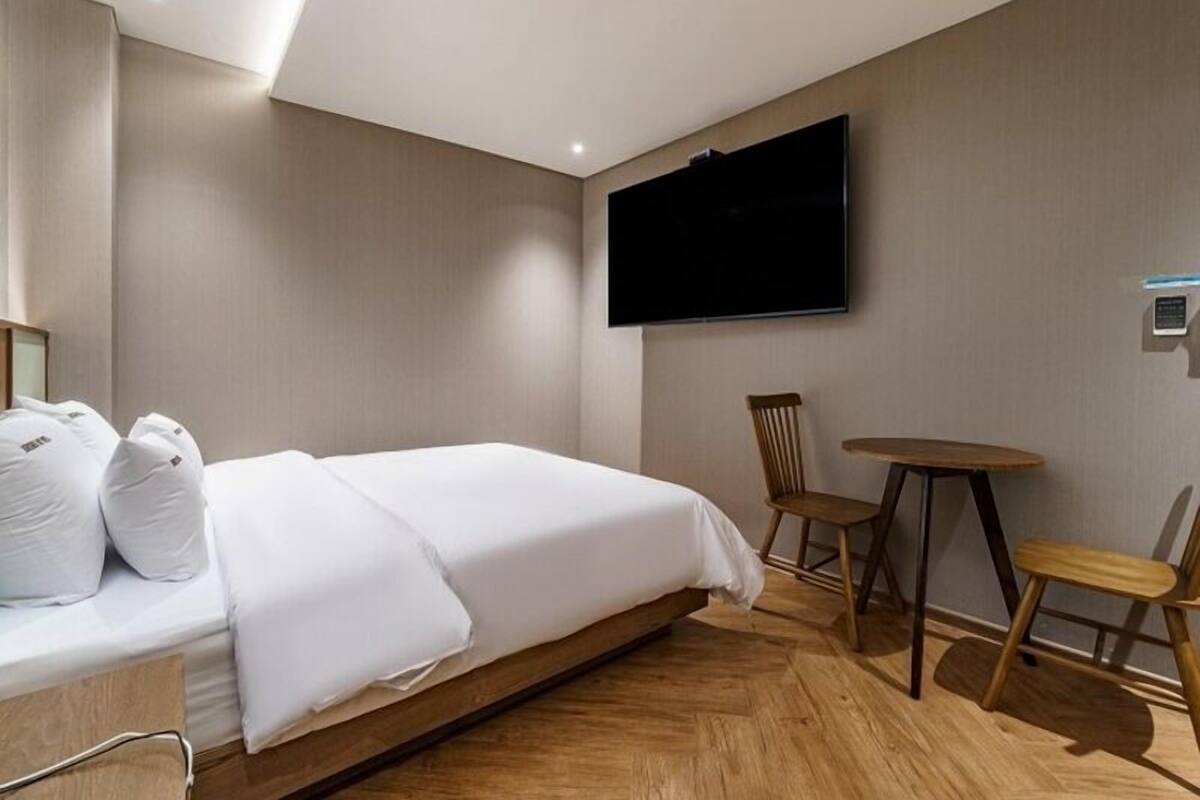 A hotel room with a bed, a flat-screen TV mounted on the wall, a wooden chair, and a small round table.