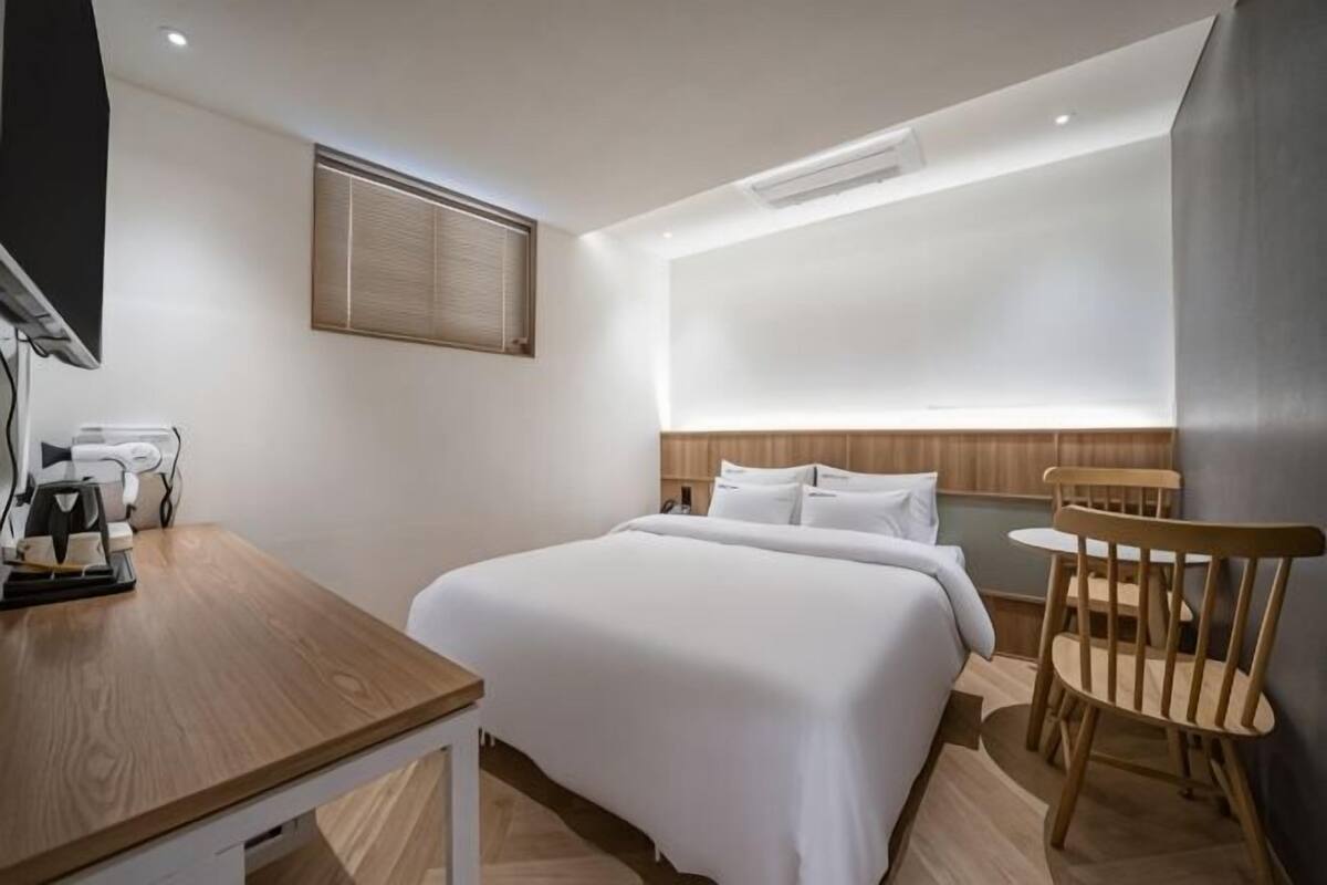 A hotel room with a bed, a desk with a chair, a small table, and a wall-mounted TV.