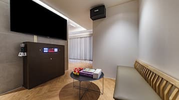 A modern hotel room with a flat-screen TV, a minibar, a small round table with books and a red bag, a bench, and a ceiling-mounted air purifier.