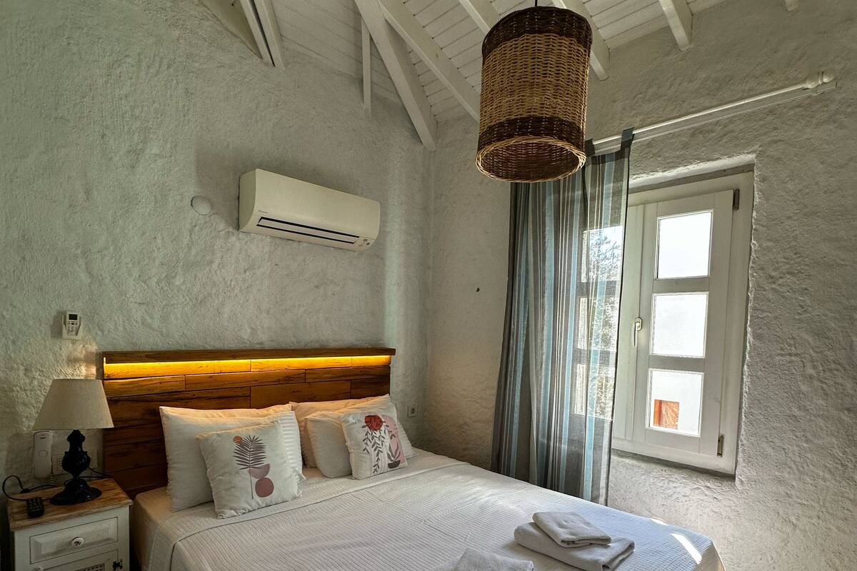 A bedroom with a wooden headboard, a large bed with white bedding, a bedside table with a lamp, an air conditioning unit, and a window with curtains.