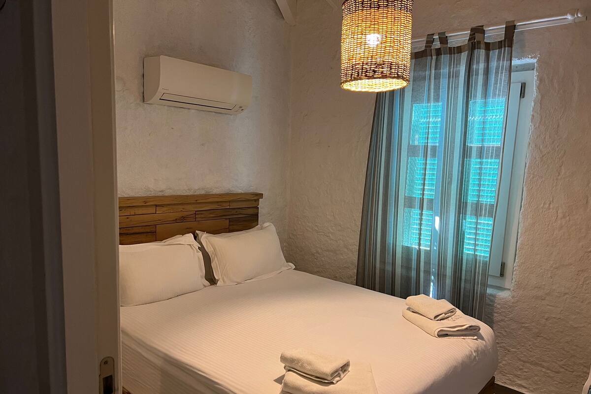 A neatly made bed with white linens and towels, a wooden headboard, a wall-mounted air conditioner, and a window with sheer curtains.