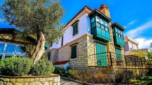 Un edificio de piedra con contraventanas verdes y techo de tejas, rodeado de vegetación y un cielo azul despejado.