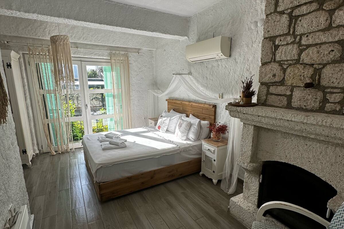 A bedroom with a wooden bed, a stone fireplace, and a view of greenery outside.