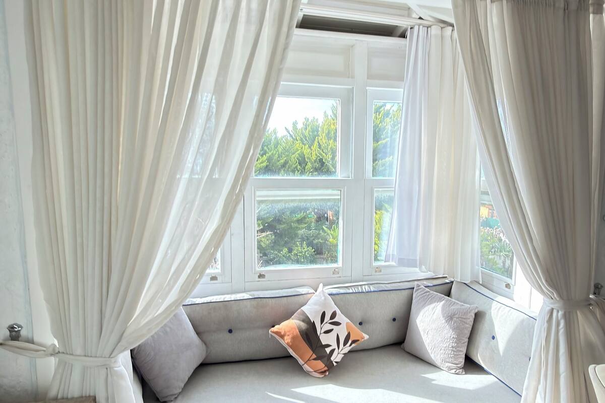 A cozy room with a built-in bench, white curtains, and a view of greenery outside.