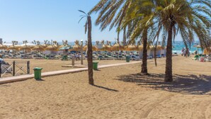 Une plage bordée de rangées de chaises longues et de parasols, avec des palmiers et un ciel dégagé.