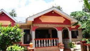A two-story house with a red and beige color scheme, a porch with a railing, and a garden with various plants.