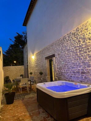 A stone-walled outdoor area with a hot tub, a table, and chairs. - Spa Workshop (Bayeux)