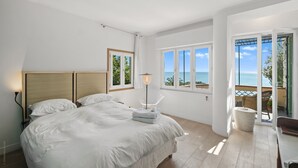 Une chambre avec un lit, une fenêtre donnant sur la mer et un balcon aménagé avec du mobilier d’extérieur.