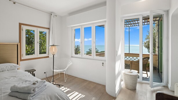 Une chambre à coucher avec un lit, une table de chevet, une lampe et une vue sur la mer grâce à de grandes fenêtres.