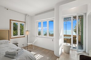 A bedroom with a bed, a bedside table, a lamp, and a view of the sea through large windows. - Exclusive Villa L'Eolienne (Nice)