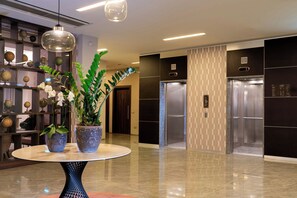A lobby with a round table, potted plants, globes on shelves, and two elevators. - Doubletree BY Hilton Milan Malpensa (SOLBIATE OLONA)