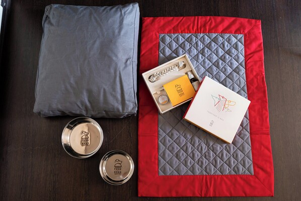 A neatly arranged set of items on a wooden surface, including a grey pillow, a red and grey quilted fabric, a small metal container with a dog logo, a yellow box with a dog logo, and a white box with a dog logo. - Doubletree BY Hilton Milan Malpensa (SOLBIATE OLONA)