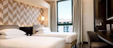 Ein Hotelzimmer mit zwei Betten, einem Schreibtisch, einem Stuhl und einem großen Fenster mit Vorhängen.