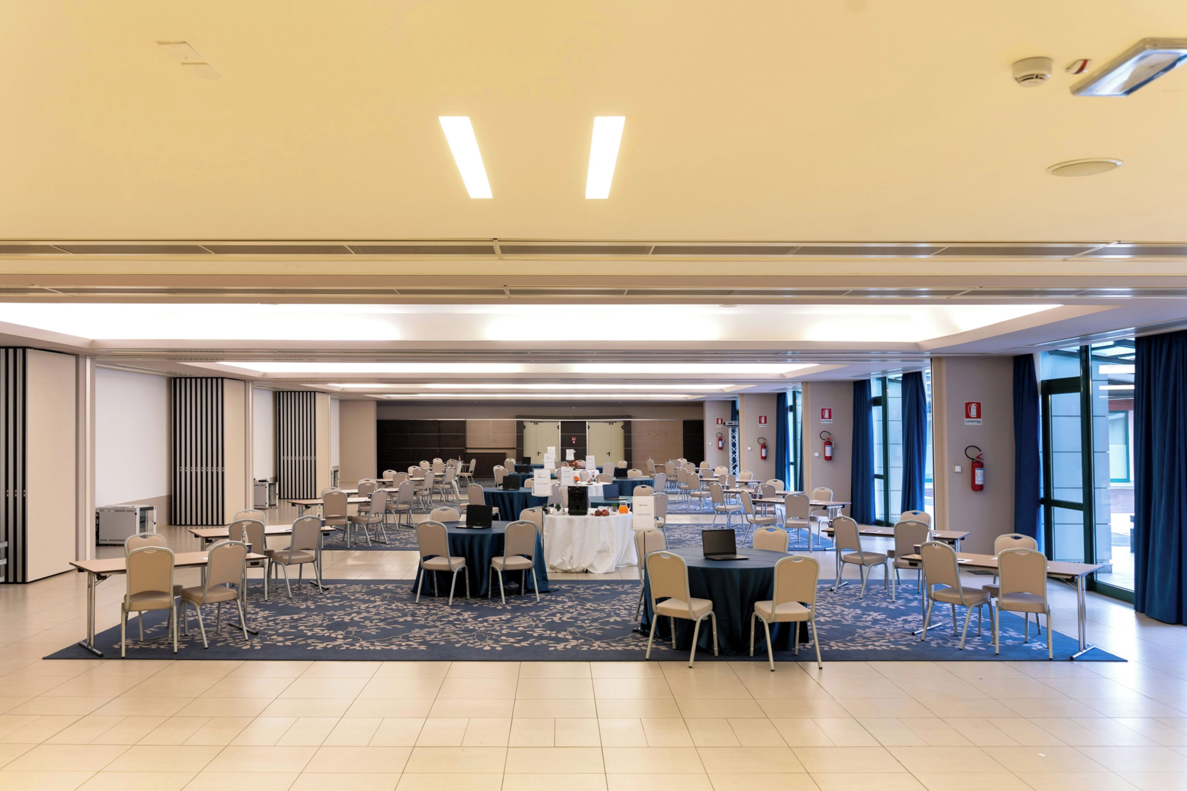 Un'ampia sala conferenze con numerosi tavoli e sedie, dotata di portatili e cartellini con i nomi, che suggerisce un contesto lavorativo o di eventi.