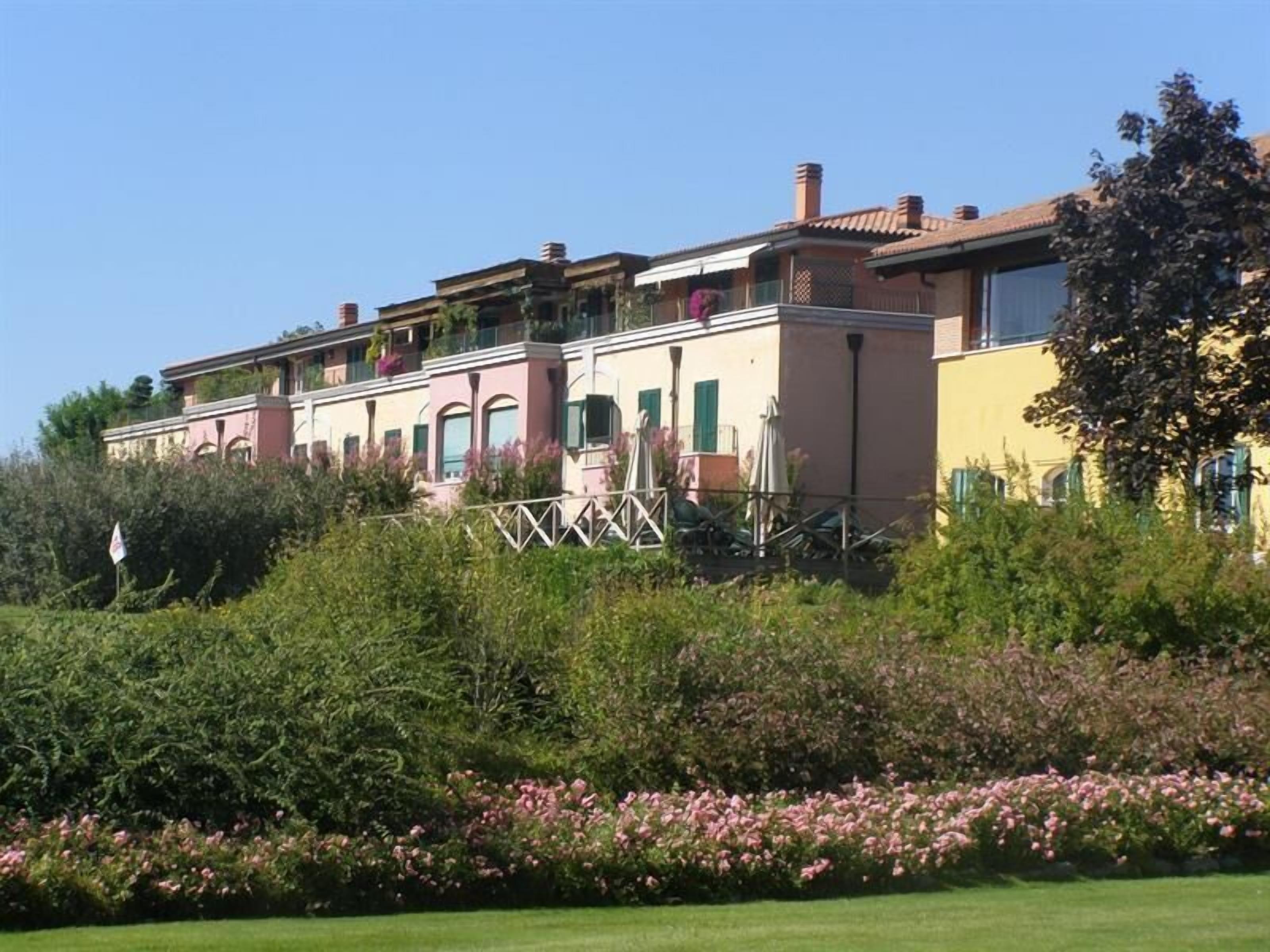 Un edificio de varios pisos con balcones, rodeado de abundante vegetación y plantas con flores.