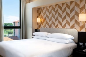 A hotel room with a large bed, two bedside lamps, a view of the outdoors, and a patterned wall. - Doubletree BY Hilton Milan Malpensa (SOLBIATE OLONA)