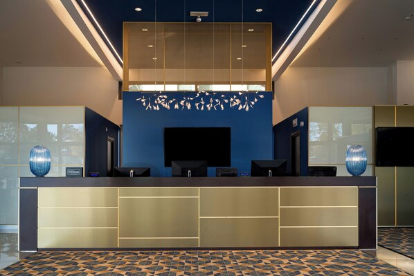 A hotel reception desk with a modern design, a large flat-screen TV, and a decorative hanging light fixture above. - Doubletree BY Hilton Milan Malpensa (SOLBIATE OLONA)