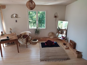 A cozy room with a wooden floor, a wicker chair, a small table with a lamp, a potted plant, and a large window with a view of greenery.