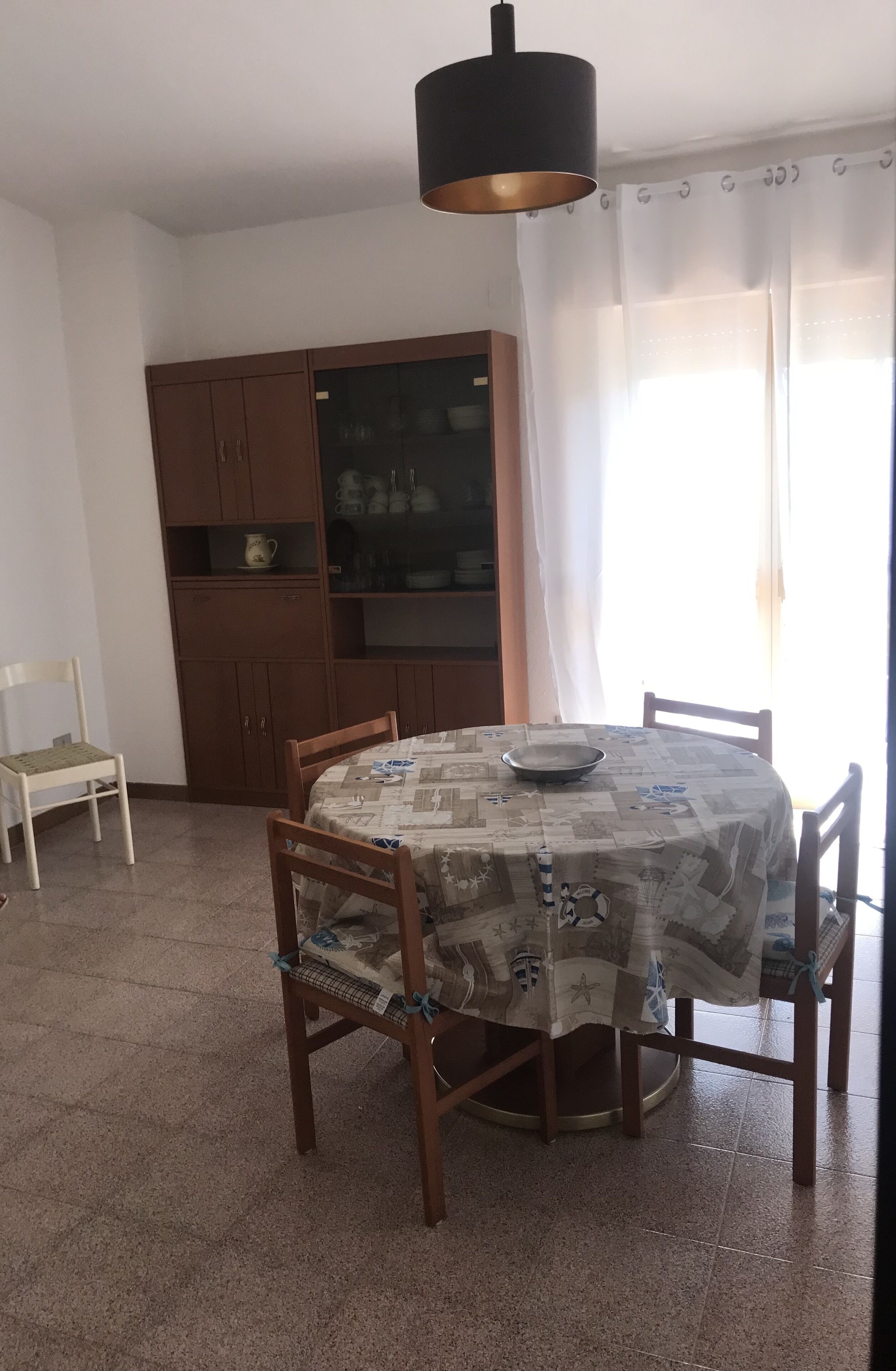 A dining area with a round table covered by a patterned tablecloth, surrounded by wooden chairs. There is a cabinet with glass doors in the background, and a white chair is placed to the left of the table.