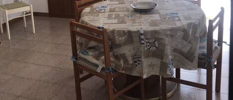 A dining area with a round table covered by a patterned tablecloth, surrounded by wooden chairs. There is a cabinet with glass doors in the background, and a white chair is placed to the left of the table.