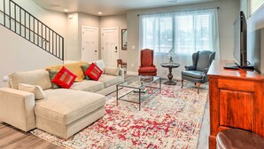 A living room with a beige sofa, red and green cushions, a glass coffee table, a patterned rug, a wooden cabinet, and a TV. - Modern Flagstaff Home, 4 Mi to Downtown! (Flagstaff)