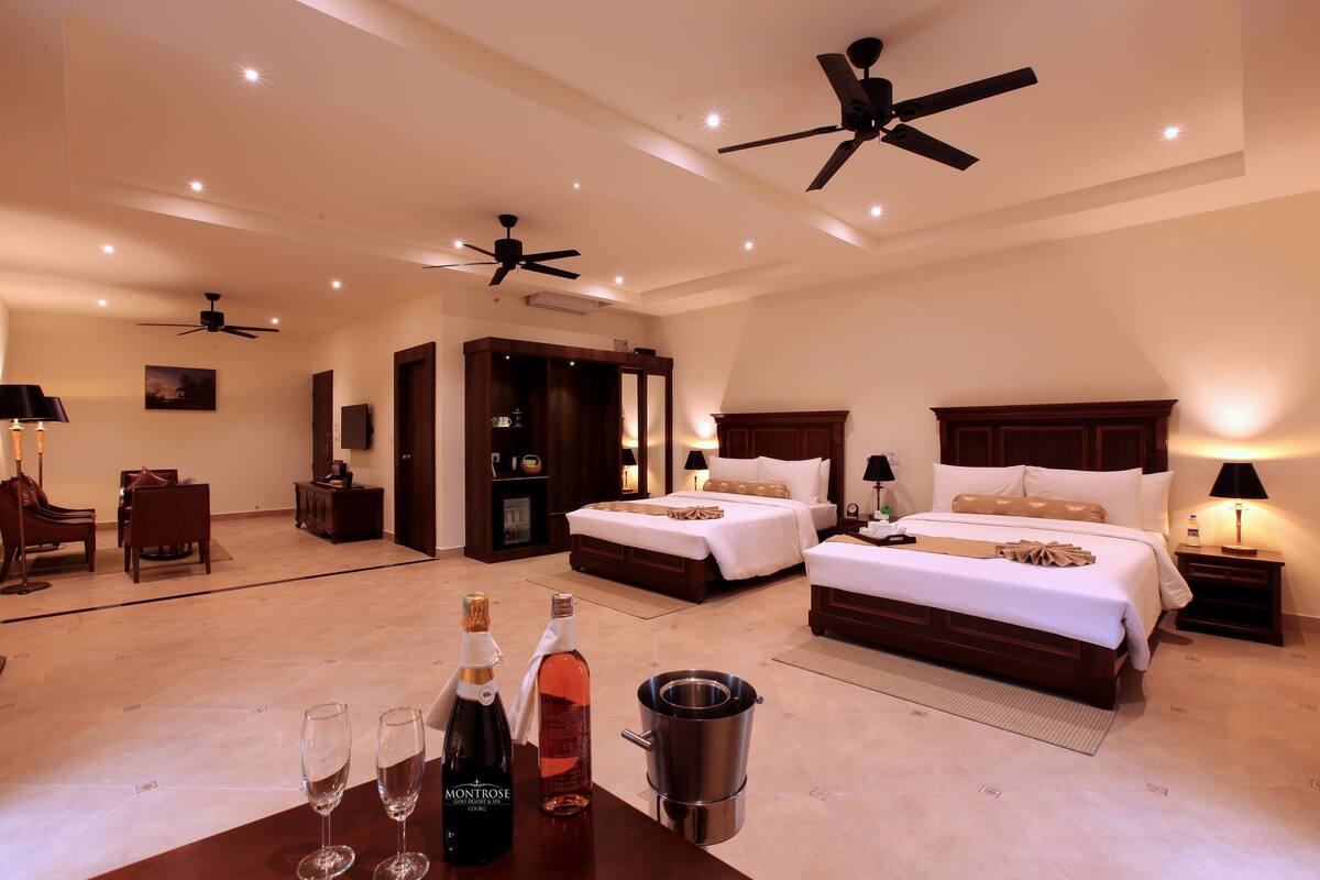 A spacious hotel room with two beds, a minibar, ceiling fans, and a dining area with a table set for two.