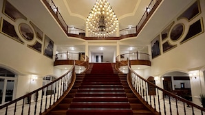 A grand staircase with a chandelier, wooden railings, and framed artwork on the walls. - Montrose Golf Resort & Spa (Somvarpet)