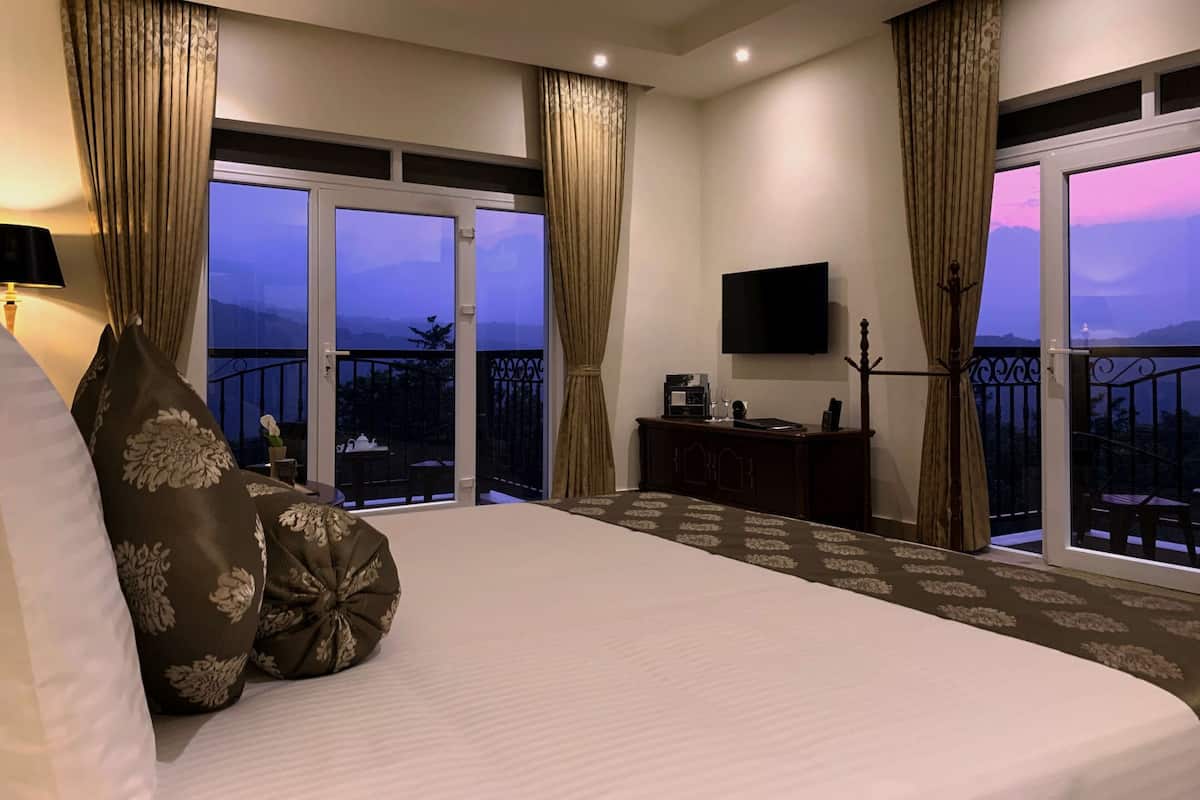 A hotel room with a large bed, a flat-screen TV, a balcony with a view of the mountains, and a desk with a coffee maker.