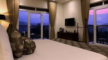 A hotel room with a large bed, a flat-screen TV, a balcony with a view of the mountains, and a desk with a coffee maker.