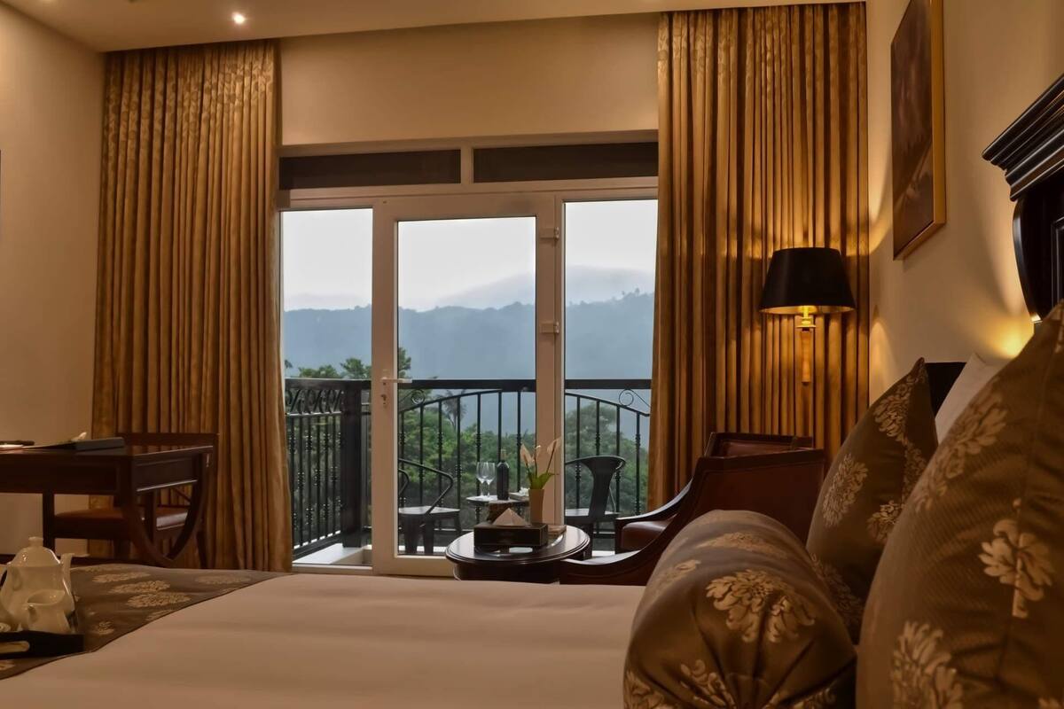 A hotel room with a bed, a desk, a chair, a balcony with a view of mountains, and a lamp.