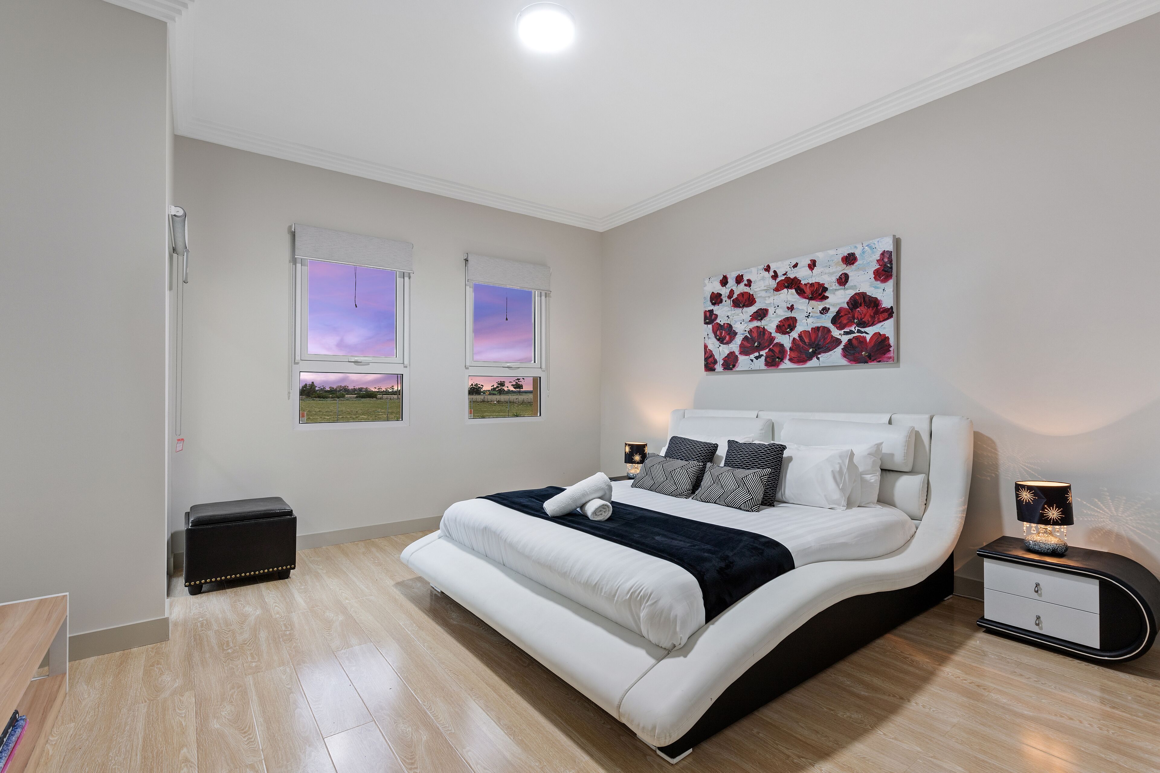 A bedroom with a large bed, two windows with scenic views, a bedside table with a lamp, and a painting on the wall.
