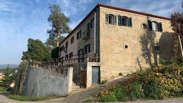 Un edificio de piedra de dos pisos con un balcón, una entrada con reja y vistas del paisaje circundante.