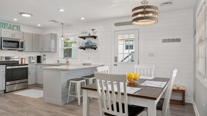 A modern kitchen with white cabinetry, a central island, and a dining area with white chairs. - Salty Pause 3 Bedroom Home in Kill Devil Hills (Kill Devil Hills)