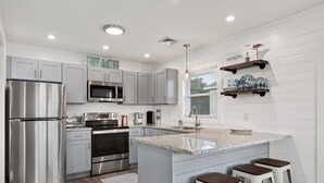 A modern kitchen with stainless steel appliances, a granite island, bar stools, and decorative shelves. - Salty Pause 3 Bedroom Home in Kill Devil Hills (Kill Devil Hills)