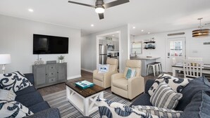 A modern living room with a sofa, armchairs, a coffee table, and a TV. There is a kitchen area with a dining table and chairs. - Salty Pause 3 Bedroom Home in Kill Devil Hills (Kill Devil Hills)
