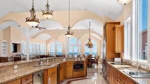 A spacious kitchen with granite countertops, wooden cabinets, and a large island. There are multiple hanging lights and a view of the ocean through large windows. - Imperial Palace Pool + Oceanfront + Pets Stay Free (Kill Devil Hills)