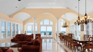 A spacious living room with a large glass table and sofas, a dining area with wooden chairs, and a view of the ocean through large windows. - Imperial Palace Pool + Oceanfront + Pets Stay Free (Kill Devil Hills)