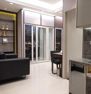 A modern living room with a sofa, a dining table with chairs, and a large sliding glass door leading to a balcony with a city view. - SUN City Apartment Sidoarjo by Group A by Zuzu (Sidoarjo)