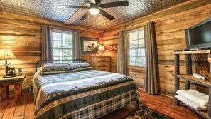 A wooden cabin bedroom with a bed, a ceiling fan, a television, and a window.