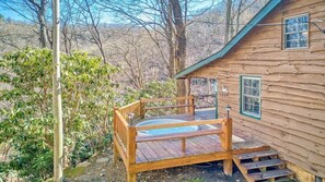 A wooden cabin with a hot tub, wooden deck, and a view of the surrounding forest.