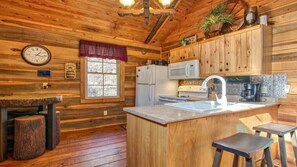 A wooden cabin interior with a kitchen area, wooden walls, a window, and a clock.