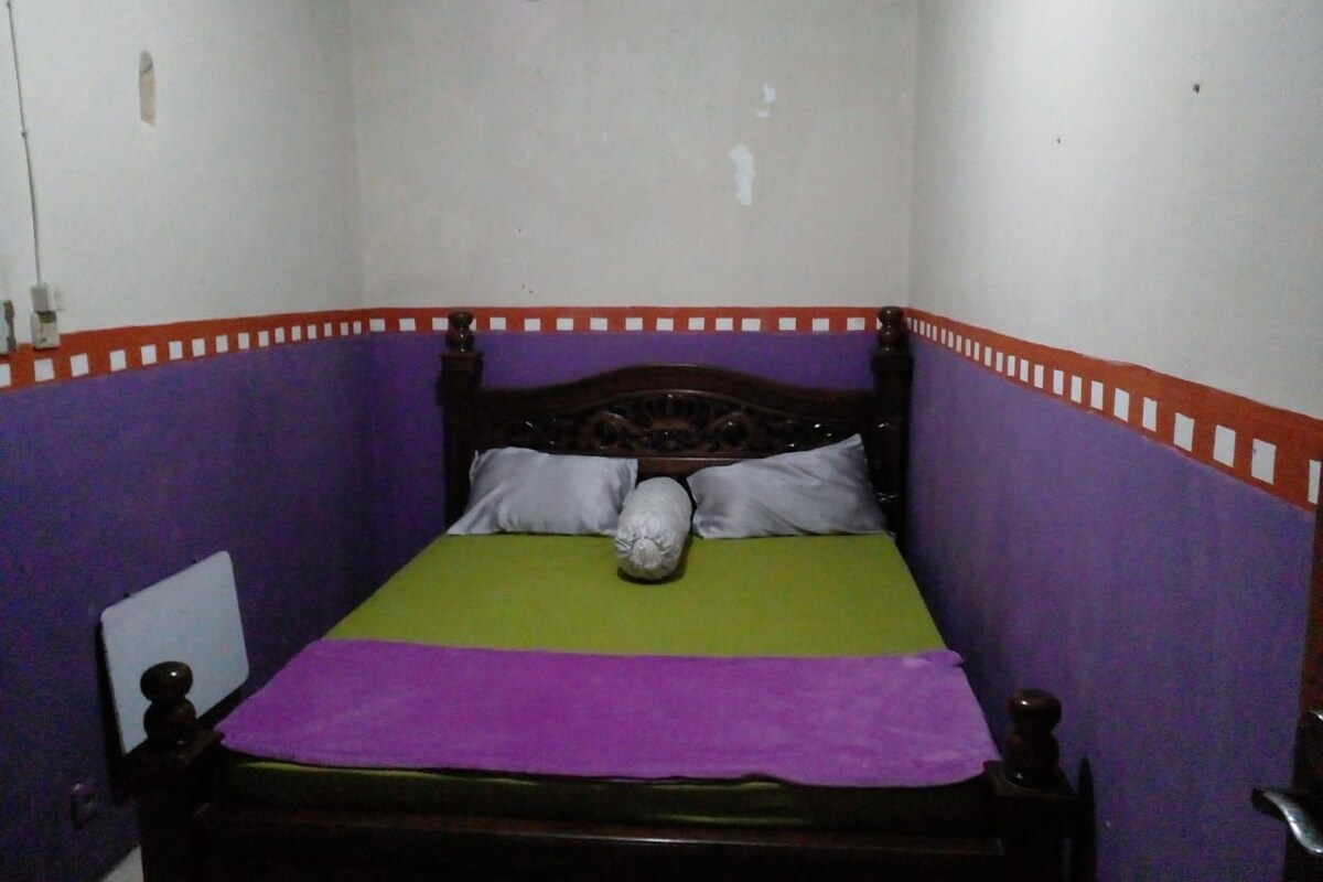 A small, single-bed room with a wooden headboard, a green and purple bedspread, and a white pillow.