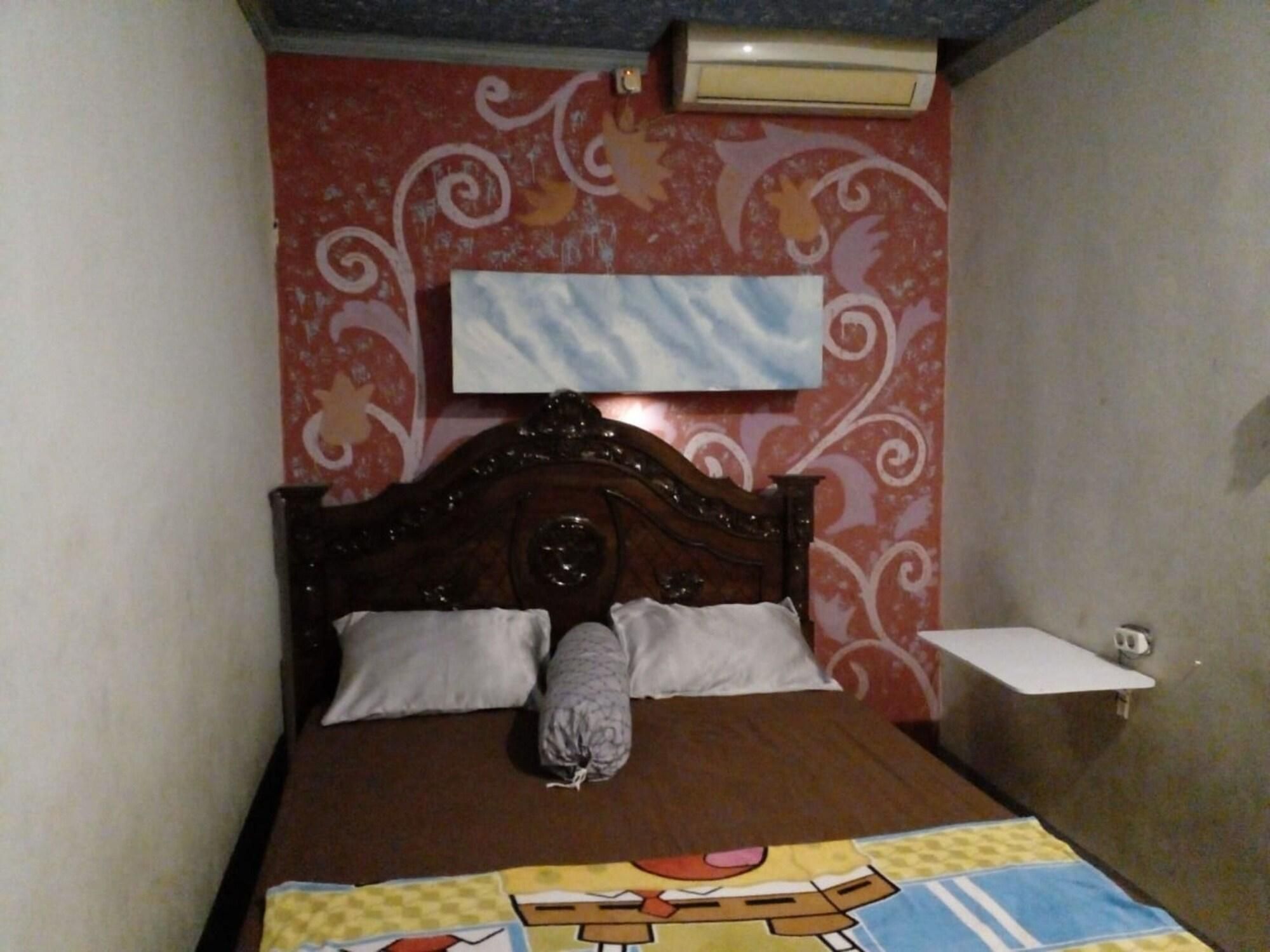 Un dormitorio con cama de madera, dos almohadas, una colcha con personajes de dibujos animados, un aire acondicionado montado en la pared y una mesita de noche pequeña.