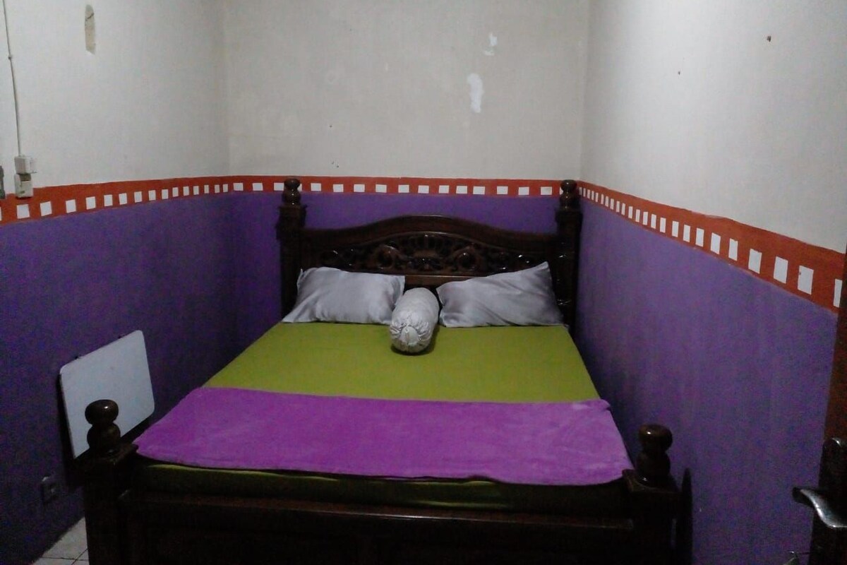 A small, narrow room with a single bed, purple walls, and a wooden headboard.