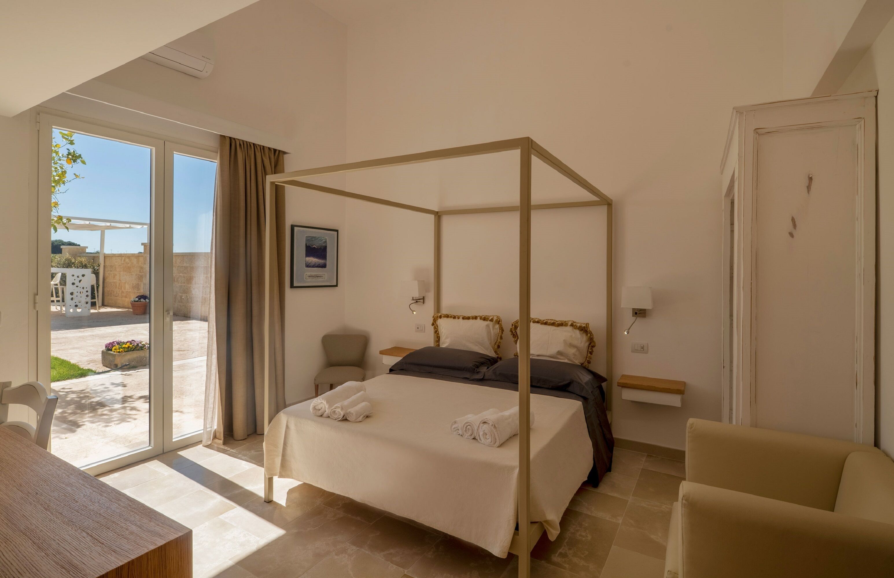 A four-poster bed with a white bedspread and two towels, a wooden nightstand, a beige armchair, and a view of the outdoor patio through a sliding glass door.